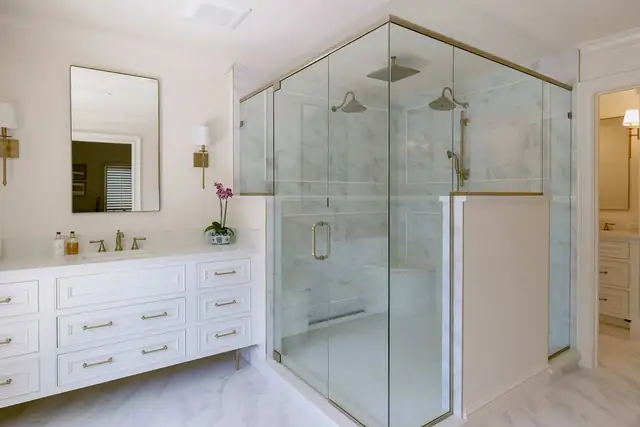 Winter Park primary bathroom with a brass-framed glass shower, marble wall tile, white double vanity and brass sconces